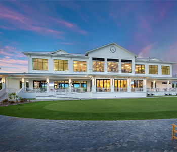 Aerial view of clubhouse beside golf course and lake with palm trees at sunset.