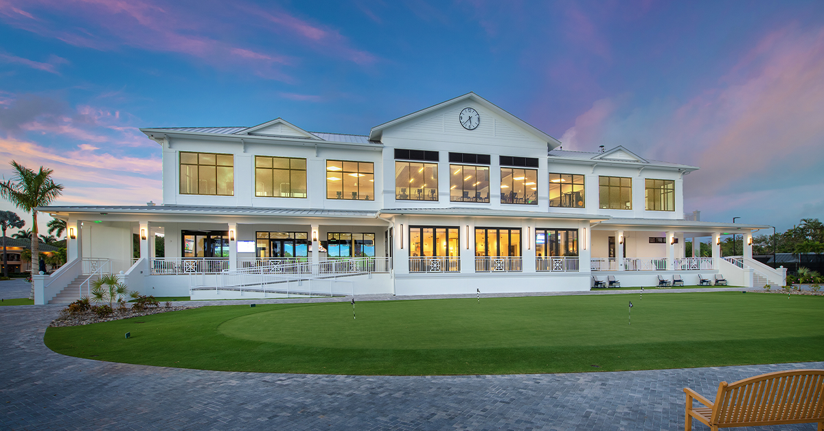 Aerial view of clubhouse beside golf course and lake with palm trees at sunset.