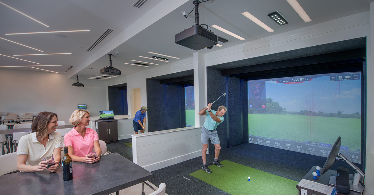 Man swinging golf club in indoor simulator lounge with large screen.