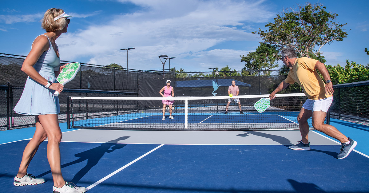Four adults playing pickleball on outdoor blue court with net and paddles.